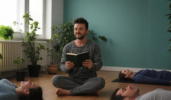 Paul Hafemann Gruppentherapie – Geführte Traumreise | Körperpsychotherapie Paul Hafemann sitzt in einem hellen Therapieraum mit türkisfarbener Wand auf einer Matte und liest entspannt aus einem dunkelgrünen Buch, während Teilnehmer mit geschlossenen Augen auf Yogamatten liegen.