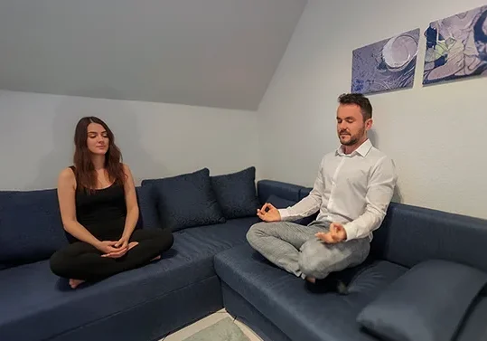 Paul Hafemann und eine Klientin sitzen barfuß im Schneidersitz auf einer dunkelblauen Couch und praktizieren gemeinsam eine meditative Atemübung; im Hintergrund sind stimmungsvolle Wandbilder zu sehen.