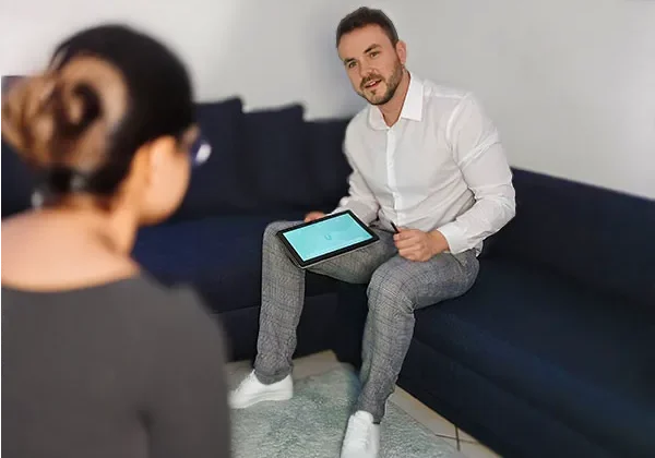 Ein männlicher Therapeut in weißem Hemd sitzt auf einer dunkelblauen Couch und hält einem Klienten ein Tablet mit schematischer Körpergrafik in grünen Akzenten entgegen; eine unscharf im Vordergrund abgebildete Person betont die Gesprächssituation.